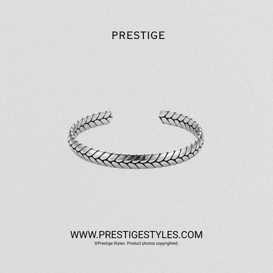 A silver-toned open cuff bracelet with a textured, woven leaf-like pattern, displayed on a light gray textured background with the brand name PRESTIGE at the top and website text at the bottom.